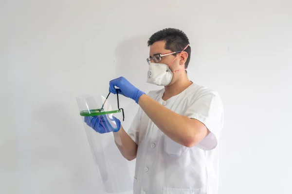 Doktor / hemşire, koronavirüsten korunmak için yüz kalkanı takıyor. O genç bir adam. Eldiven, maske ve gözlük takıyor. Beyaz bir üniforma giyiyor..