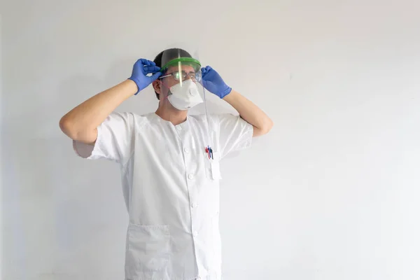 Doktor / hemşire, koronavirüsten korunmak için yüz kalkanı takıyor. O genç bir adam. Eldiven, maske ve gözlük takıyor. Beyaz bir üniforma giyiyor..