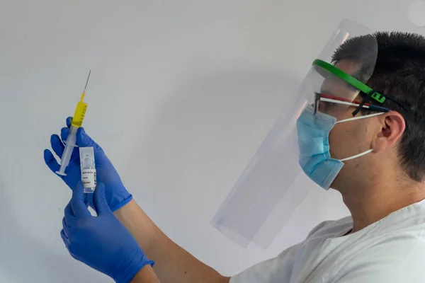 Doctor with face shield shows a vaccine. In his hand he has the vaccine and a syringe. He wears a white uniform, wears a mask, gloves and glasses.
