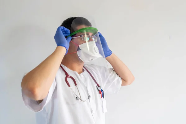 Male nurse putting on a face shield He is a young man. He wears gloves, masks, and stethoscope. He wears a white uniform.