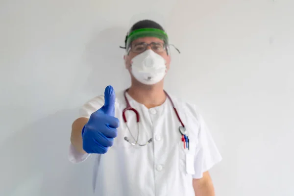 Yüz kalkanı olan bir erkek hemşire. O genç bir adam. Eldiven, maske ve steteskop takıyor. Beyaz bir üniforma giyiyor..