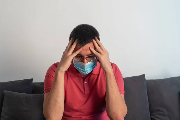 Endişeli adam. Kafasına eliyle dokunuyor, stresli görünüyor. Maske, polo tişört ve gözlük takıyor. Kanepede oturuyor..
