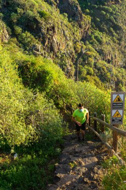 Koşu antrenmanı yapan adam. Maskesi var. Adam ormanda bir yolda koşuyor. Heyelan ve kaygan zemin nedeniyle bir tehlike işareti görülebilir..