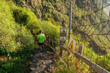 Koşu antrenmanı yapan adam. Maskesi var. Adam ormanda bir yolda koşuyor. Heyelan ve kaygan zemin nedeniyle bir tehlike işareti görülebilir..