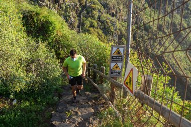 Koşu antrenmanı yapan adam. Bir maskesi ve bir şişe suyu var (yumuşak matara). Adam ormanda bir yolda koşuyor. Heyelan ve kaygan zemin nedeniyle bir tehlike işareti görülebilir..