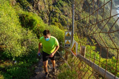 Koşu antrenmanı yapan adam. Bir maskesi ve bir şişe suyu var (yumuşak matara). Adam ormanda bir yolda koşuyor. Heyelan ve kaygan zemin nedeniyle bir tehlike işareti görülebilir..