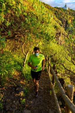 Patikadan aşağı koşan maskeli bir adam. Koridor bir ormandadır. Fotoğraf Tenerife 'de çekildi. İdeal bir iz sürme yeri. Elinde bir şişe su var (Yumuşak matara)).