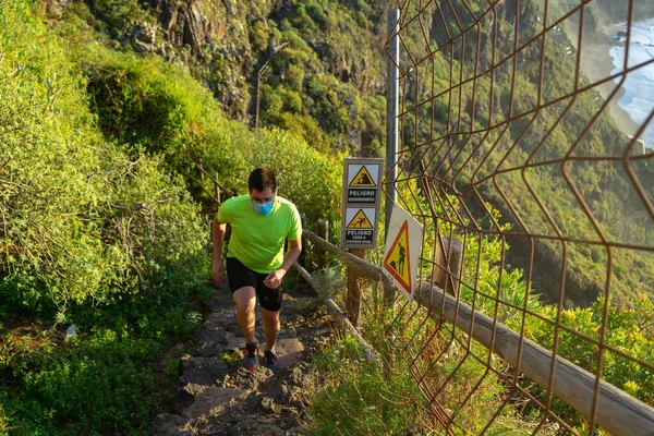 Koşu antrenmanı yapan adam. Maskesi var. Adam ormanda bir yolda koşuyor. Heyelan ve kaygan zemin nedeniyle bir tehlike işareti görülebilir..