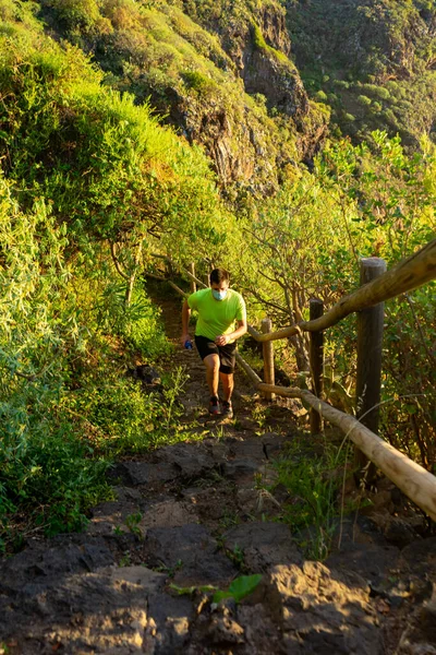 Man with mask running down a path. The corridor is in a forest. The photo was taken in Tenerife, an ideal place to practice trail running. In his hand he has a bottle of water (Soft flask).