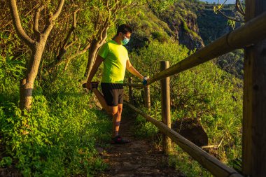 Koşucu bacağın kuadriseps kaslarını geriyor. Adam bir ormanda ve tahta korkuluklara yaslanıyor. Elinde bir maske ve yumuşak bir matara var..
