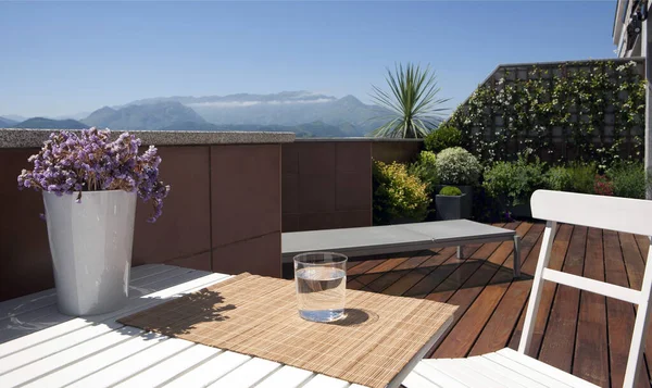 Terraza soleada con mesa, silla, vaso de agua y plantas y vistas a una montaa