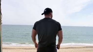 Adam deniz kenarında, kumsalda antrenman yapıyor, denize ve kuma bakıyor, boks yapıyor, koşuyor, telefonda konuşuyor, deniz taşı fırlatıyor.