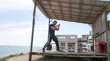 Adam deniz kenarında, kumsalda antrenman yapıyor, denize ve kuma bakıyor, boks yapıyor, koşuyor, telefonda konuşuyor, deniz taşı fırlatıyor.