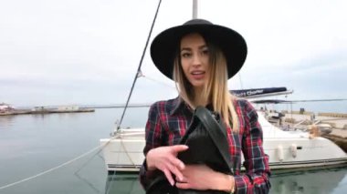 Limanda, yatların yanında poz veren kız, kısa bir elbise, yuvarlak bir şapka ve yüksek çizmelerle yürüyor, gülümsüyor.