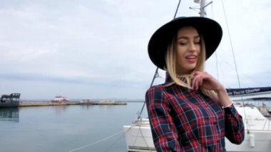 Limanda, yatların yanında poz veren kız, kısa bir elbise, yuvarlak bir şapka ve yüksek çizmelerle yürüyor, gülümsüyor.