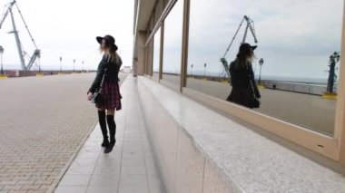 Limanda, yatların yanında poz veren kız, kısa bir elbise, yuvarlak bir şapka ve yüksek çizmelerle yürüyor, gülümsüyor.