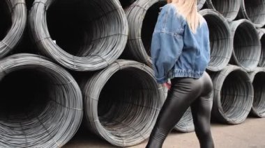 Metal bir kabloya yakın poz veren, efsanevi beyaz ayakkabılar giyen, kot ceket giyen, dans eden, gülümseyen bir kız.