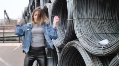 Metal bir kabloya yakın poz veren, efsanevi beyaz ayakkabılar giyen, kot ceket giyen, dans eden, gülümseyen bir kız.