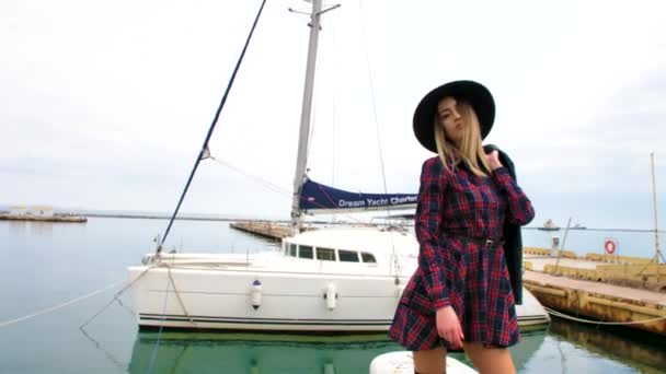 女孩在海港靠近游艇的地方摆姿势 身穿短裙 头戴圆帽 脚穿高跟鞋 面带微笑 — 图库视频影像