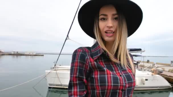 女孩在海港靠近游艇的地方摆姿势 身穿短裙 头戴圆帽 脚穿高跟鞋 面带微笑 — 图库视频影像