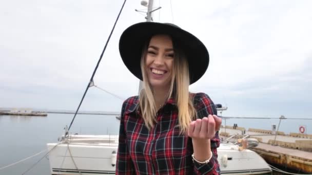 ヨットの近くの海港にポーズをとっている女の子は 短いドレス 丸い帽子と高いブーツで歩き 笑っています — ストック動画