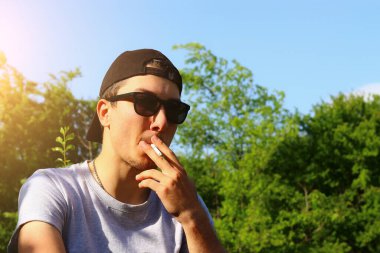 Siyah tişörtlü ve güneş gözlüklü bir adam, başlık takıyor, bulanık bir arka planda sigara içiyor, metin ya da yazıt koymak için fotokopi alanı var, tonlanmış..