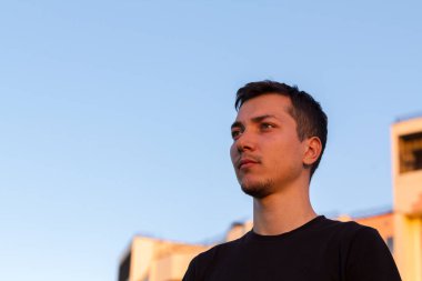 Siyah tişörtlü bir adam şehrin bulanık arka planında sabahın erken saatlerinde düşünceli bir şekilde mesafeye bakar. Alt görünüm