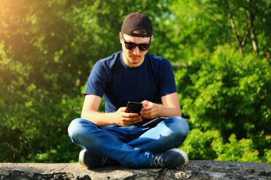 Sıradan giyinen sıradan bir adam, temiz havada akıllı telefonuyla oturuyor ve sosyal ağlarda mesajlaşıyor.