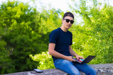 Freelancer, bir şehir nehrinin beton çitinde otururken dizüstü bilgisayarla çalışıyor.