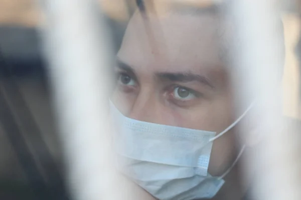 Sad young man in medical mask on quarantine. Window with bars and reflection, toned