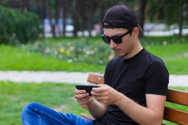 Genç bir adam ya da erkek, öğrenci ya da serbest çalışan, şehir parkında bir bankta otururken dinleniyor. İş ve eğlence için konforlu şehir ortamı