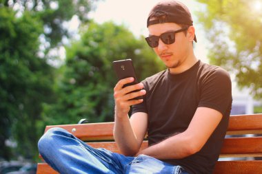 Genç bir adam şehirde bir bankta oturuyor ve akıllı telefonun ekranına bakıyor. Ayarlanmış