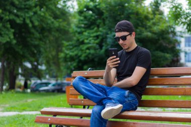 Genç bir adam ya da erkek, öğrenci ya da serbest çalışan, şehir parkında bir bankta otururken dinleniyor. İş ve eğlence için konforlu şehir ortamı