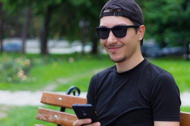 Genç bir adam ya da erkek, öğrenci ya da serbest çalışan, şehir parkında bir bankta otururken dinleniyor. İş ve eğlence için konforlu şehir ortamı