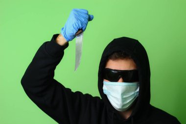 Tıbbi maske takmış genç bir gangster ya da suçlu siyah kapüşonlu güneş gözlüğü takmış elinde bir bıçak tutuyor.