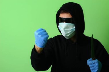 Tıbbi maske takmış genç bir gangster ya da suçlu siyah kapüşonlu güneş gözlüğü takmış elinde bir bıçak tutuyor.