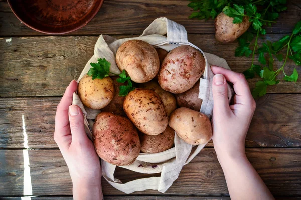 Fresh raw potatoes in a linen bag on a wooden background. Top view. Free space for text. Woman holds potatoes in a hand