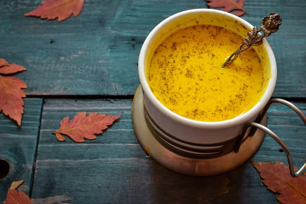 Golden milk in a white cup. Wooden color blue background. Autumn leaves. A healthy drink made from milk, turmeric, cinnamon and honey. Yellow milk