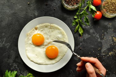 Kadın kahvaltıda yumurta yiyor, çatal tutuyor. Tabakta pişmiş yumurta. Üst manzara, koyu arkaplan. Kahvaltı ve malzemeler..