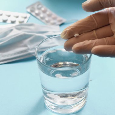 Cerrahi maskeli ve eldivenli Coronavirus profilaksi. Tıbbi maske, haplar, uyuşturucular, bardaktaki su. Açık mavi arkaplan. Metin için boş alan. Eldivenli eller.