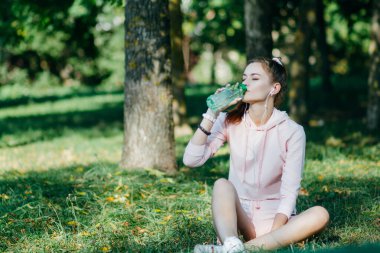 Genç kadın, koşudan sonra su içerek ve çimlerde oturarak dinleniyor.