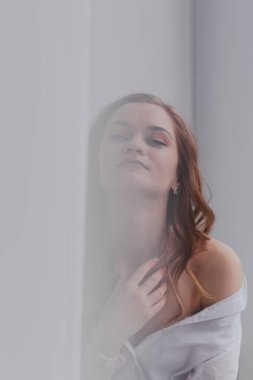 Genç, uzun saçlı, beyaz gömlekli, pencere eşiğinde oturan ve perdelerin arasından kameraya bakan bir esmer.