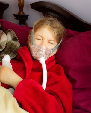 Kırmızı bornoz giymiş bir kadın, solunum maskesi takmasını gerektiren bir hastalıktan kurtulurken yatakta uyuyor. Belli ki yoğun bakım gerektiren bir solunum sorunundan etkilenmiş..