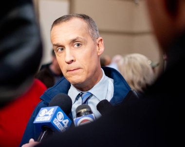 Corey Lewandowski, Bedford New Hampshire 'da düzenlenen GOP 2020 kutlamaları sırasında gazetecilerin sorularını sıralarken basının gürültüsüyle çevrelenmişti. Mavi bir pire ceketi, mavi gömlek ve mavi kravat giyiyor. Kırmızılı bir kadın saate odaklanmış.