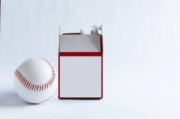 A baseball rest next to its box with some of the paper wrapping escaping from the top.