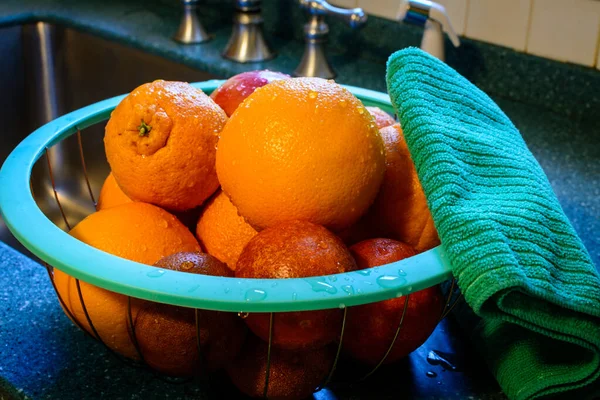 Freshly cleaned basket of citrus fruit rests in a basket on the kitchen counter next to stainless steel sink in a kitchen. Water droplets cling to the fruit signify that it was just sanitized. Next to the basket of fruit is a fluffy green towel.