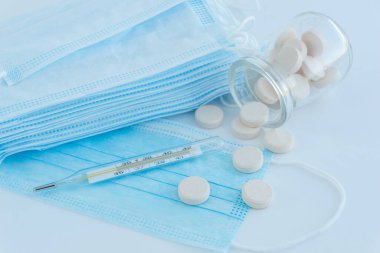 Mavi tıbbi maske yığını, açık mavi arka plan, cıva termometre, virüs salgınlarından ve koronavirüsten korunmak için geniş açık sarı tabletli cam bir kavanozun yanında yer alır.