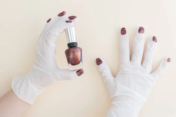 hand in medical glove with red nails holds nail polish on light background, nurse wants to be beautiful during coronavirus pandemic, making beauty with closed nail salons