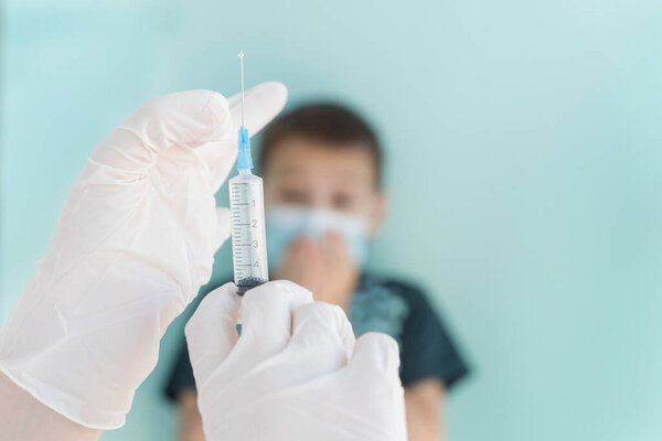 hands in white medical gloves hold syringe with medicine in it, prepare for injection of vaccine, child in medical mask on background not in focus look with fear, scared of vaccination from viruses