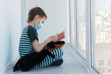 Pijama giyen ve köpek oyuncağıyla tıbbi maske takan okul çocuğu, apartman balkonu, uzaktan eğitim, ev okulu konsepti, karantina ve sels-tecrit üzerine kitap okuyor. 